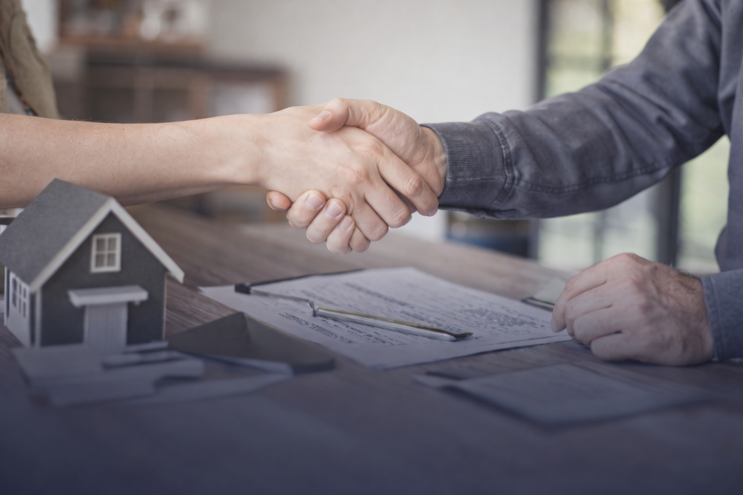 Homeowner and roofing professional shaking hands with insurance paperwork and a model house on the table