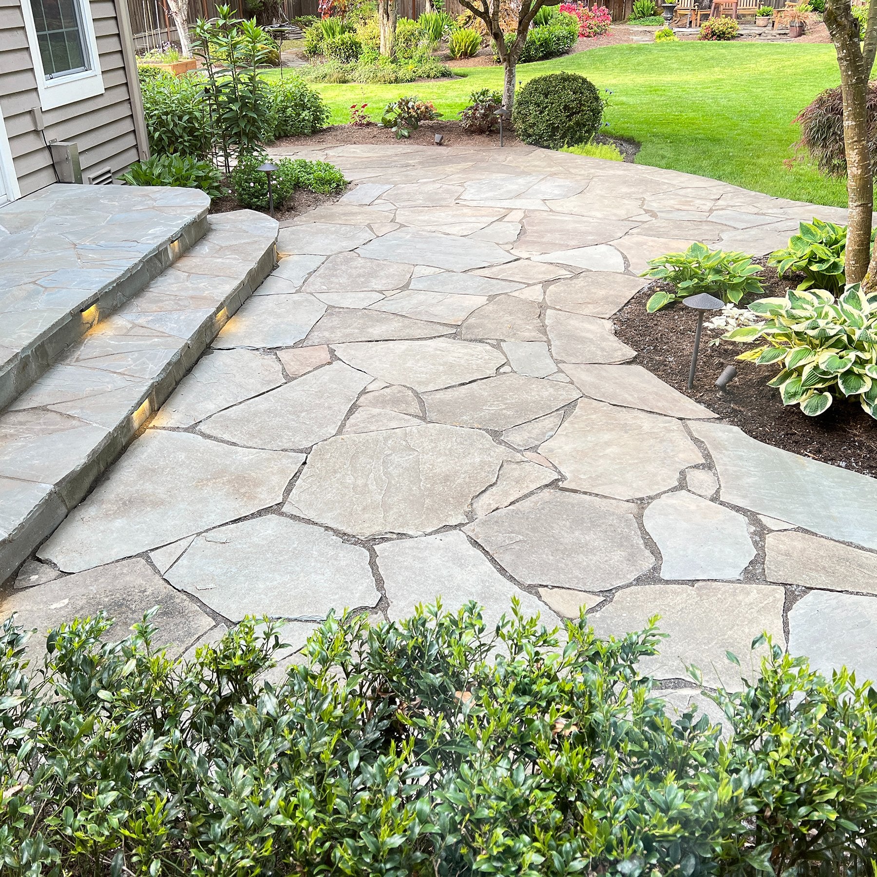 Backyard flagstone patio with stone steps, garden lighting, and landscaped greenery around a residential home.