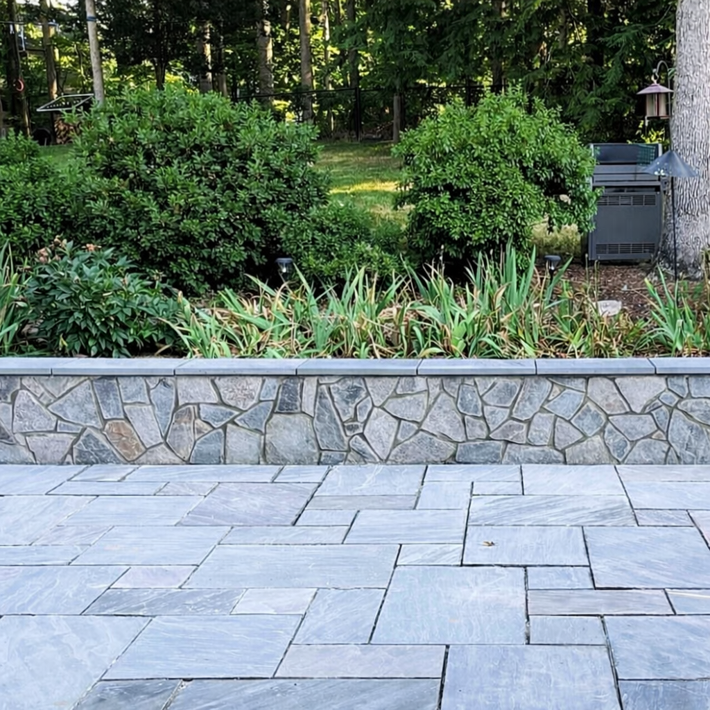 Stone retaining wall with paved walkway and landscaped plants in a residential backyard.